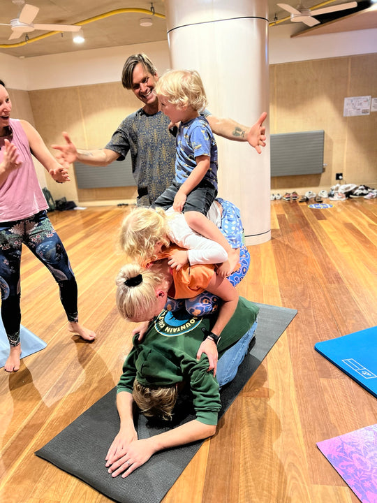 Children and adults stacking into a playful Rainbow Yoga group shape with Gopala smiling nearby